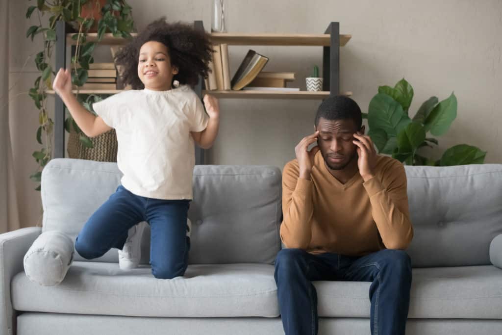 Dad sitting on couch with ADHD daughter jumping and yelling next to him.