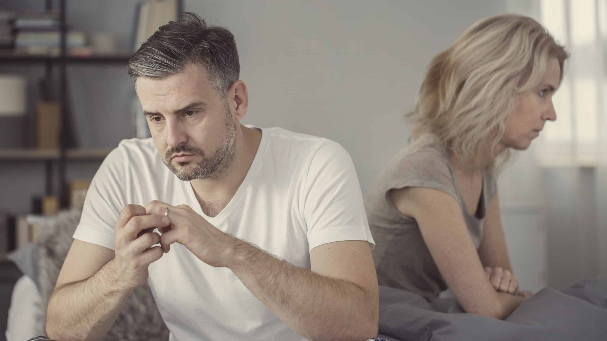 Depressed middle-age couple seated in different directions. He’s toying with his wedding band, implying a troubled marriage