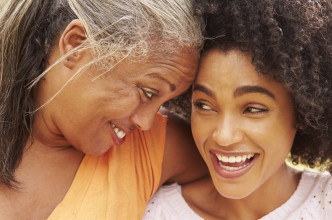 Smiling black mom side hugging her laughing adult daughter, resting her forehead against her daughter’s