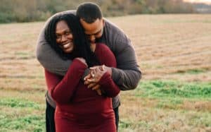 A photo of Jason brown hugging his wife, Tay, from behind. They are standing outdoors in a field.