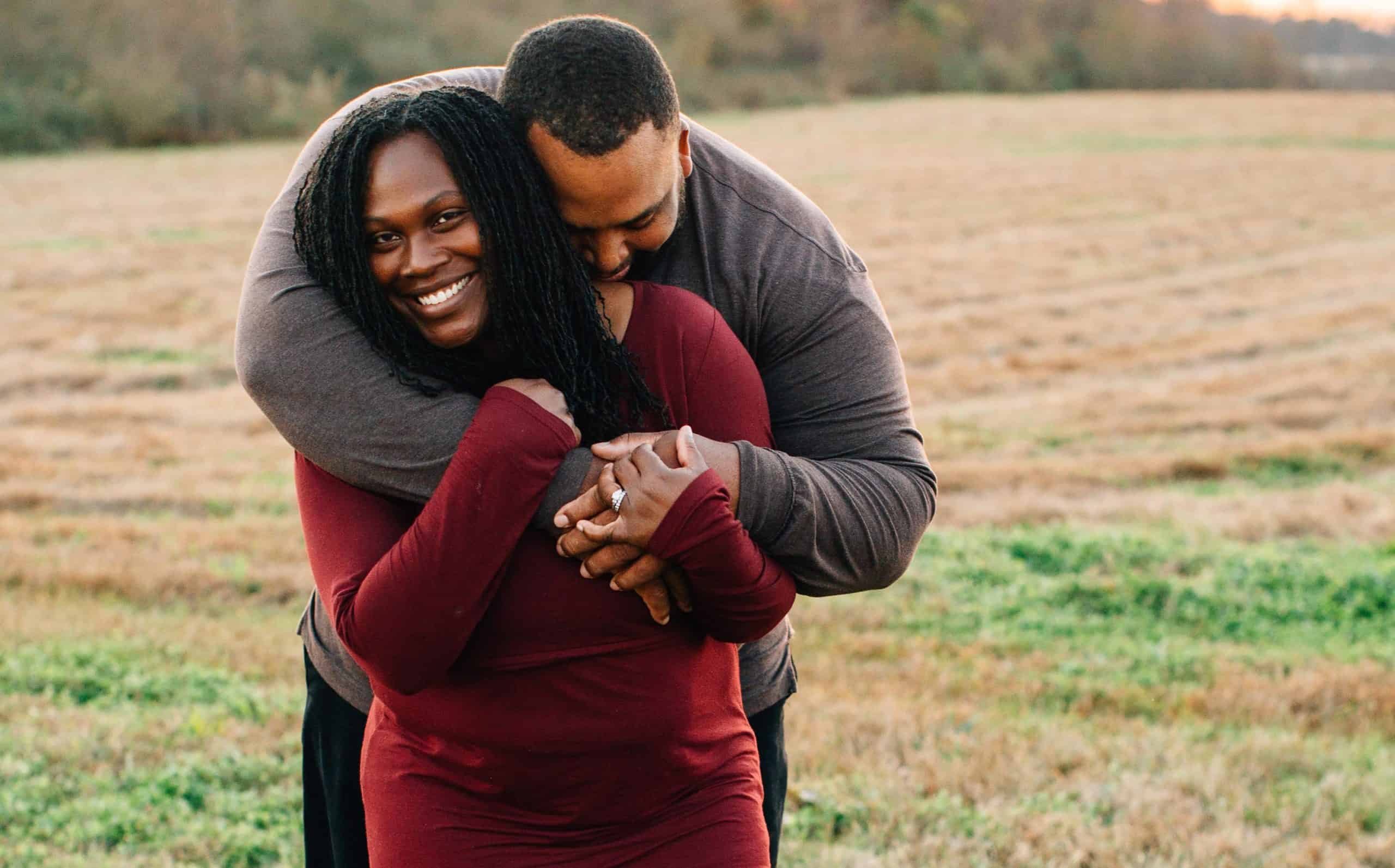 A photo of Jason brown hugging his wife, Tay, from behind. They are standing outdoors in a field.