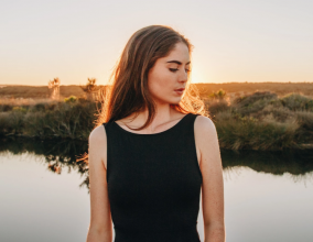 Young woman in a black dress in front of a calm stream, looking off to the side pensively