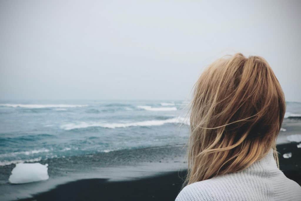 Shown from behind, a woman standing on a beach, looking out over the ocean on a cloudy day