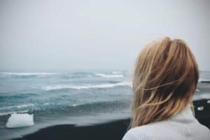 Shown from behind, a woman standing on a beach, looking out over the ocean on a cloudy day