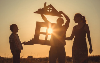 Silhouette of mom and young son holding cardboard cutout of incomplete house while dad attaches the roof