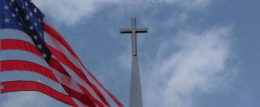Cross atop a church steeple with backdrop of partly cloudly sky and U.S. flag waving in the foreground