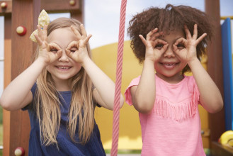 Young black girl and white girl smiling, holding okay hand gestures to their eyes, looking through them like binoculars