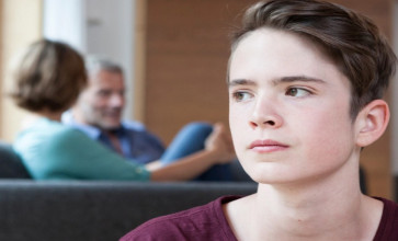 Pensive, gender-ambiguous teen in foreground, parents sitting on couch having discussion in background