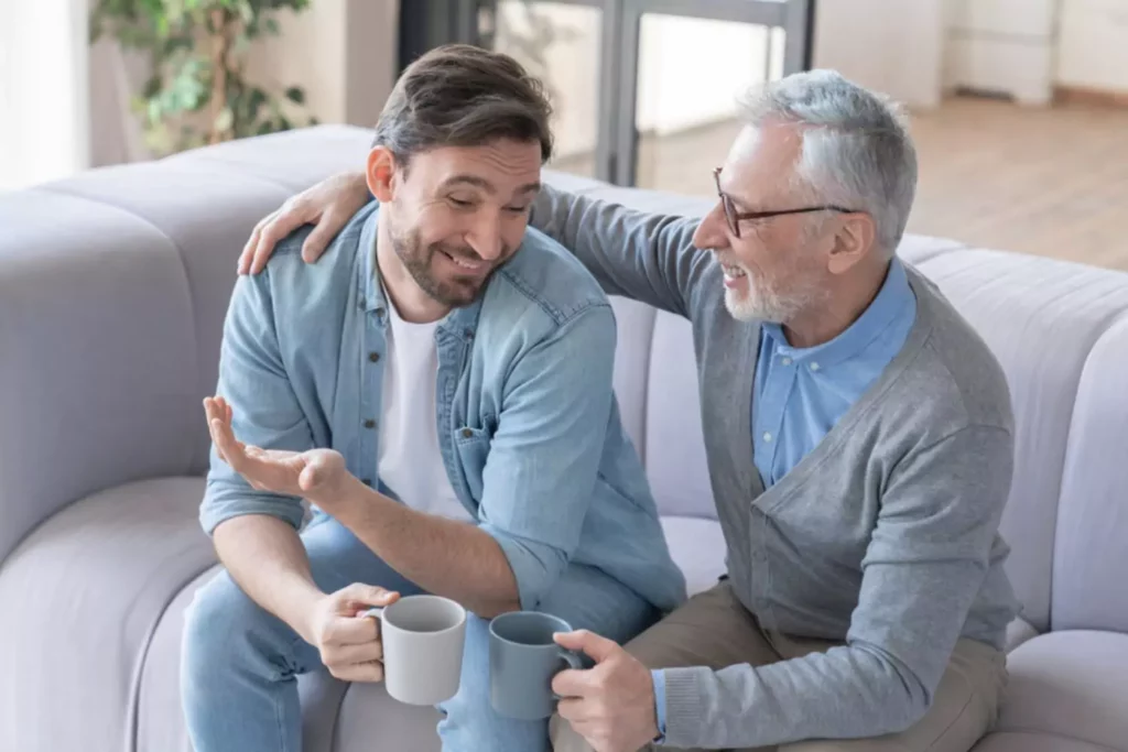 setting boundaries with adult children can be good. This graying father and his son are sitting on the couch smiling and talking