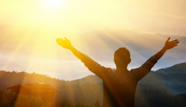 Shown from behind, a silhouette of a young man standing on a hill raising his arms toward the sunrise over the mountains