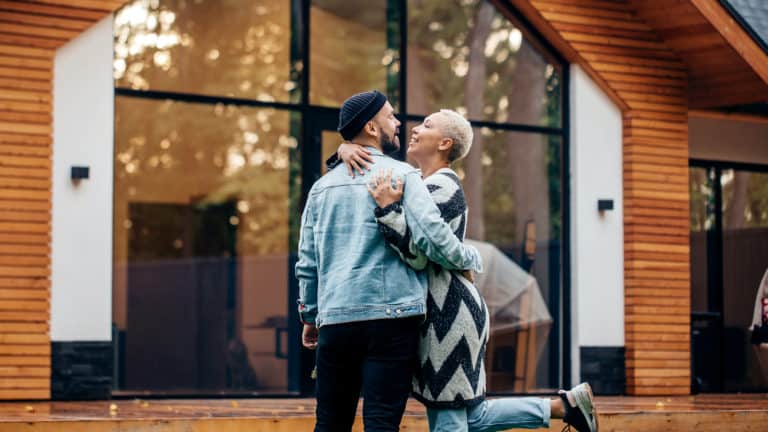 A happy couple embraces in front of their modern home, symbolizing the choice between investing in a wedding day or prioritizing marriage and a mortgage.