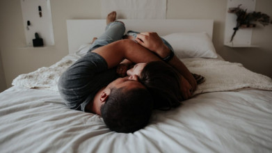 Clothed couple lying together in bed; he's holding her in his arms