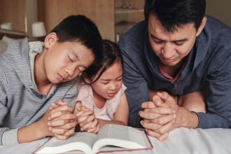 Father and his two young children praying together at a bedside with an open bible in front of them