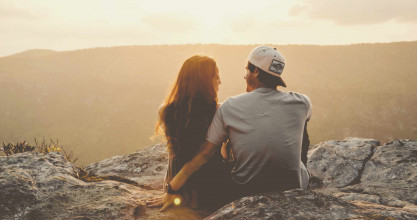 Shown from behind, a smiling couple looking at each other while sitting on a hilltop with a mountain view
