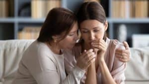 Middle-age woman comforting and side-hugging a young, worried woman as they sit on a couch