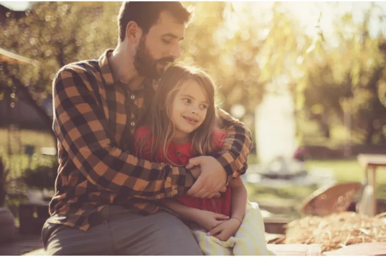 Father with an arm around his young daughter as they sit outdoors on a bench