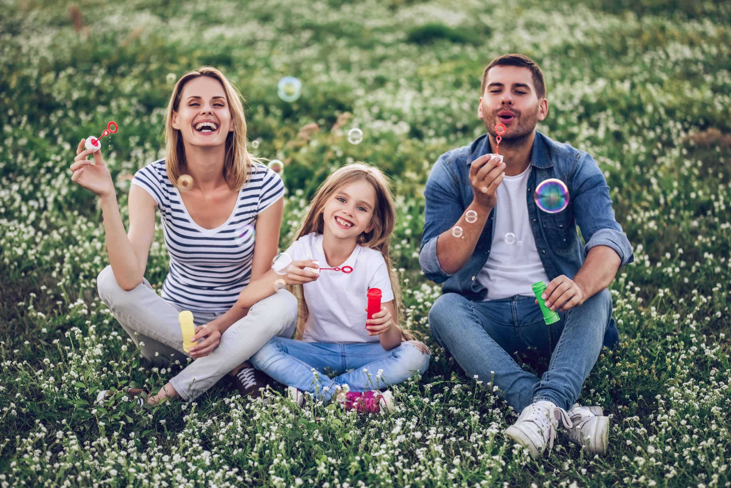 Happy mom, dad, and young daughter sitting in a field and blowing bubbles