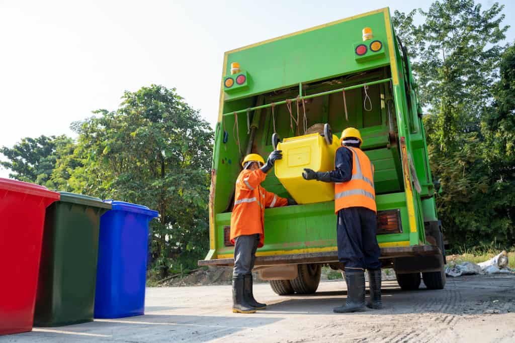 Two sanitary workers dump a trash can into a garbage truck.