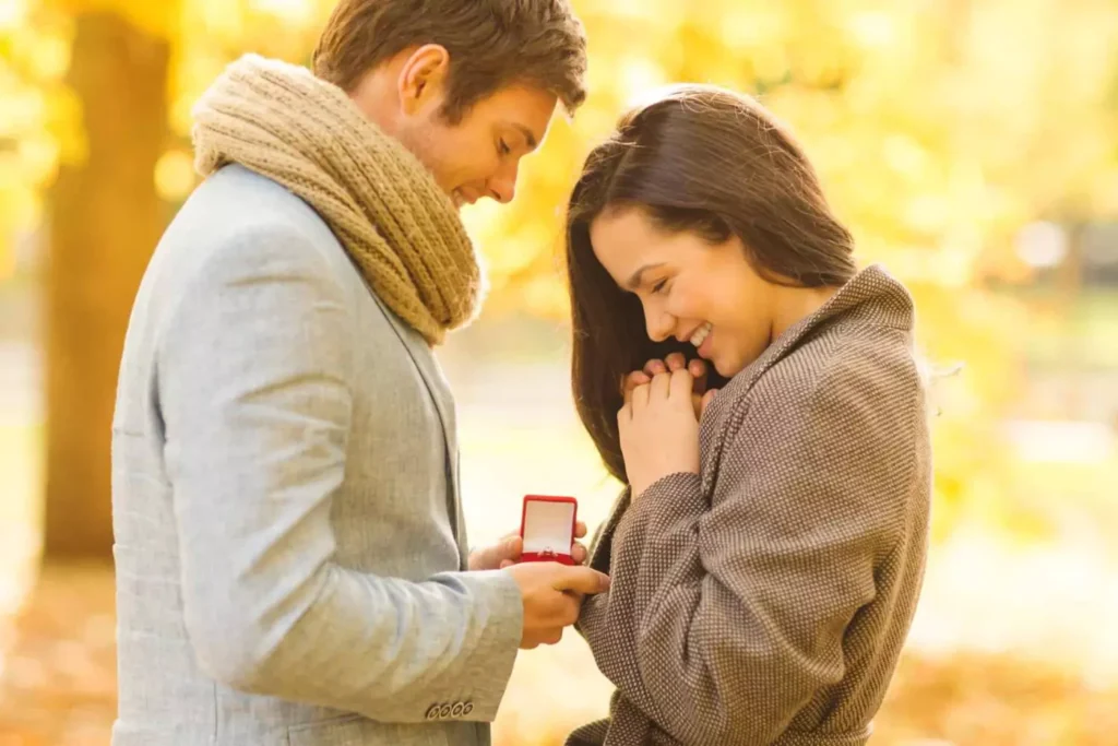 A young couple shares a joyful moment as the man presents an engagement ring in a small red box, symbolizing the “ring by spring” tradition.