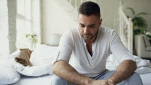 a man in white shirt sits on bed thinking with woman sleeping in background wondering should men have say in abortion