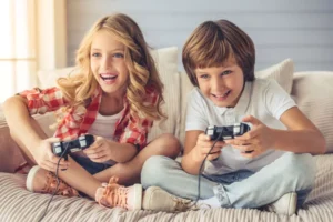 When to play video games? Two kids, a brother and sister about eight years old sitting on a couch playing video games. Both kids have a big smile on their face.