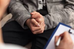 A man in counseling with his hands clasped, and counselor listening with pad at the ready.