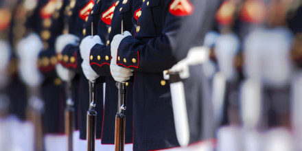 Shown from waist level, a row of US Marines standing in formation in their dress uniforms, holding their rifles