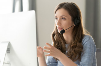 Woman in headphones with microphone consulting client on phone in customer support service, looking at computer screen