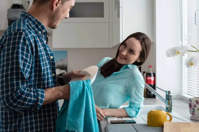 A newlywed couple shares a loving moment in the kitchen as the husband washes dishes, representing the need for good marriage advice for the first year of marriage—building connection through everyday moments.