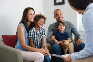 Happy young family sitting on couch and talking with family counselor. Smiling parents with adopted children discussing with counselor. Multiethnic family meeting a financial agent.