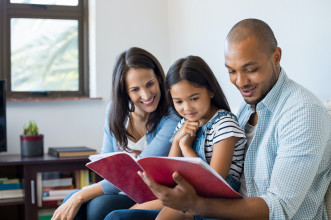 Happy multiethnic family sitting on sofa together looking at daughter's drawing book. Black father reading the homework on exercise book of his cute kid. Parents feeling proud on progress of daughter.
