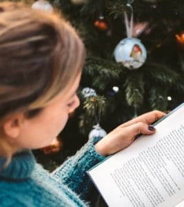 A young woman sitting next to a Christmas tree reading a book