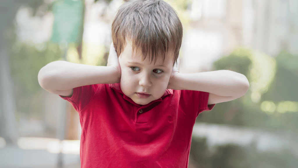 An autistic little boy in a red shirt covering his ears. One of the possible signs of autisim.