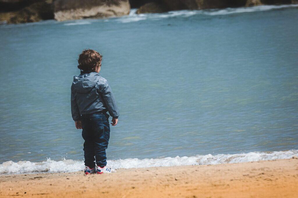 back of young boy standing on beach looking at water