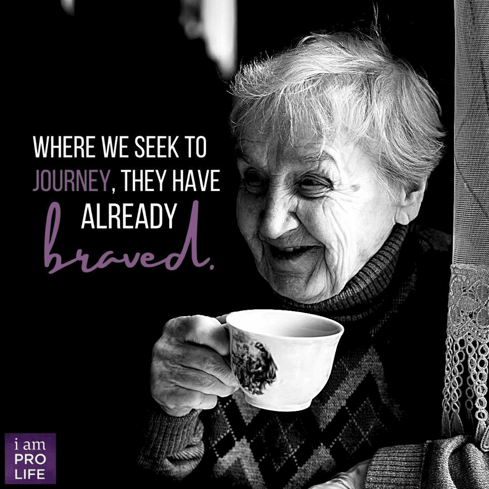 An elderly woman sips a cup of tea, happy to interact with those who are cartaking for the elderly.