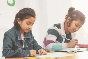 Two elementary school girls sit side by side at a wooden table, focused on drawing and writing with colored pencils and markers.