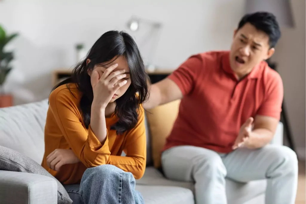 A distressed woman sits with her head in her hand while a man angrily gestures towards her, depicting emotional abuse in marriage.
