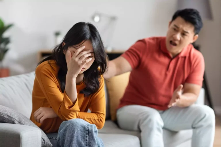 A distressed woman sits with her head in her hand while a man angrily gestures towards her, depicting emotional abuse in marriage.