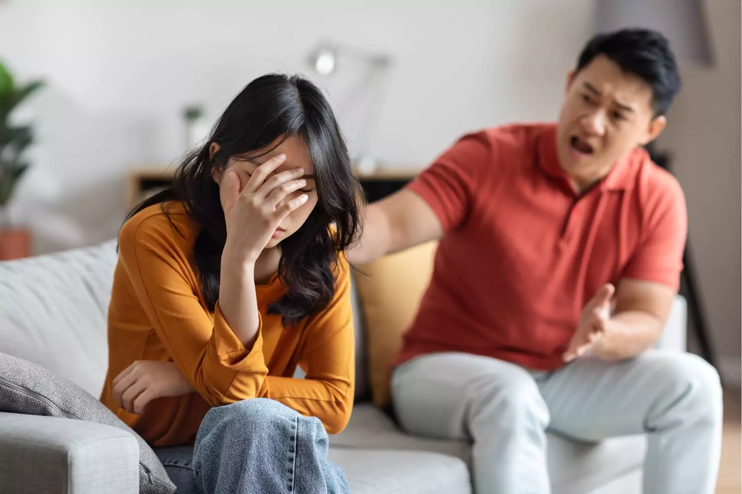 A distressed woman sits with her head in her hand while a man angrily gestures towards her, depicting emotional abuse in marriage.