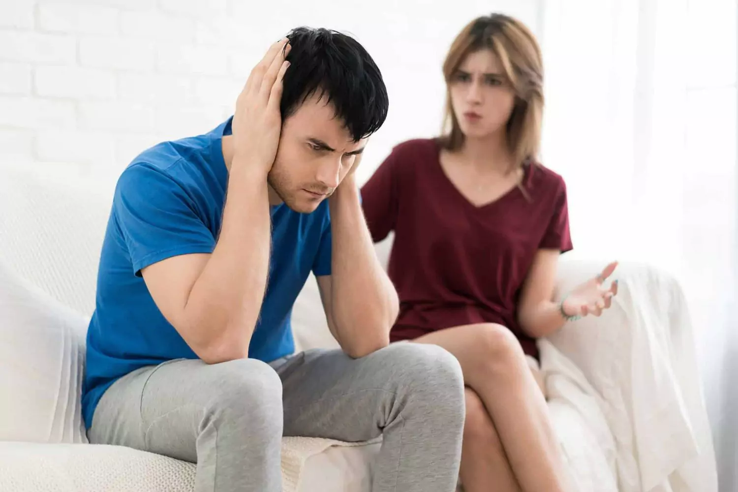 A frustrated man sits on a couch covering his ears while a woman, appearing upset, gestures at him — illustrating the feeling of a husband saying, “My wife constantly criticizes me.”