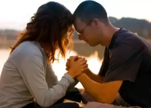 Couple praqying together as a couple with hands clasped, sitting by a peaceful lakeside at sunset, symbolizing spiritual intimacy and connection