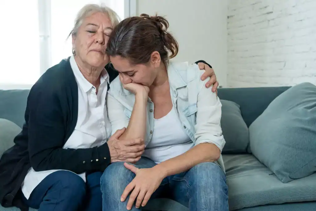 Two individuals sit close together on a couch, with one person leaning forward and the other placing an arm around them in a supportive posture, illustrating the theme of coping with the loss of a child to suicide.