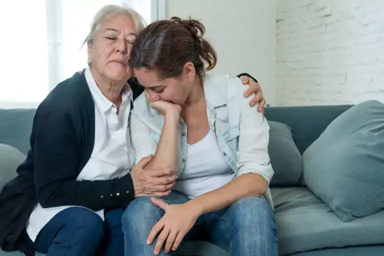 Two individuals sit close together on a couch, with one person leaning forward and the other placing an arm around them in a supportive posture, illustrating the theme of coping with the loss of a child to suicide.