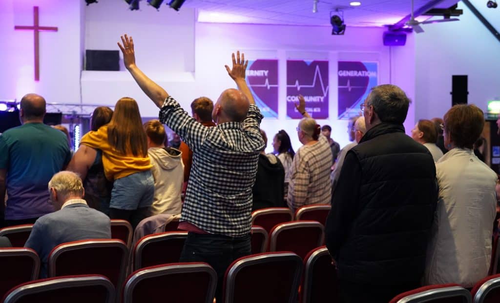 Photo of a man worshipping in Church, arms outstretched, with other people in the seats.