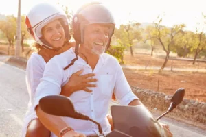 A joyful couple rides a scooter down a scenic road, sharing laughter with their spouse as they embrace the adventure together.