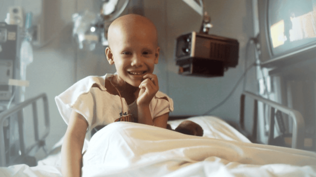 Little girl with childhood cancer, bald, sitting up in a hospital bed smiling