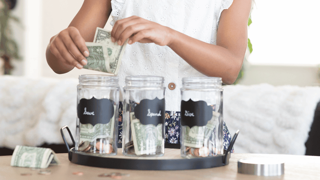 Photo of a girl holding money over three different jars labeled "give" "save" "spend"