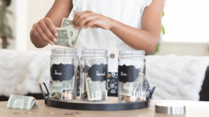 Photo of a girl holding money over three different jars labeled "give" "save" "spend"