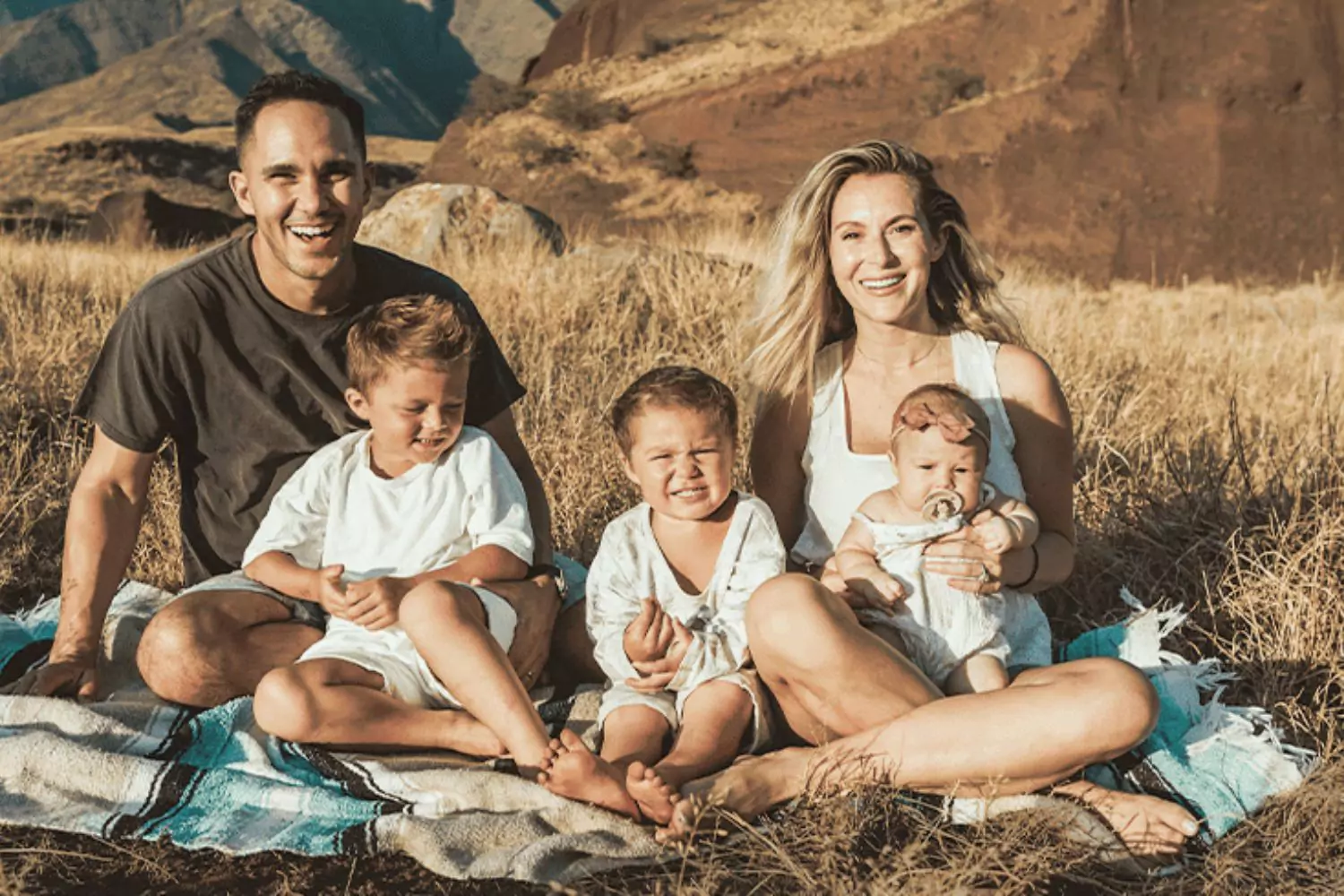 Carlos and Alexa PenaVega sit on a picnic blanket with their three young children, smiling in a scenic outdoor setting.