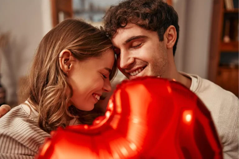 A couple smiling warmly with their foreheads touching, holding a red heart-shaped balloon, exemplifying how to have a happy Valentine’s Day with your spouse through shared affection and joy.